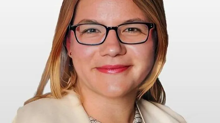 A woman with glasses, blonde hair and a light-coloured jacket smiles at the camera.