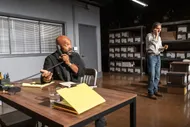 Two men talk on phones in an office filled with shelves of labelled boxes.